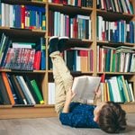 Boy laying on  the floor with the feet up, reading a book against multi colored bookshelf in library. Education, Knowledge, Bookstore, Lecture. Pupil holds a book in his hands.