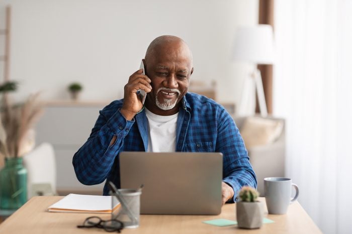 Mature African Man Talking On Cellphone Using Laptop In Office